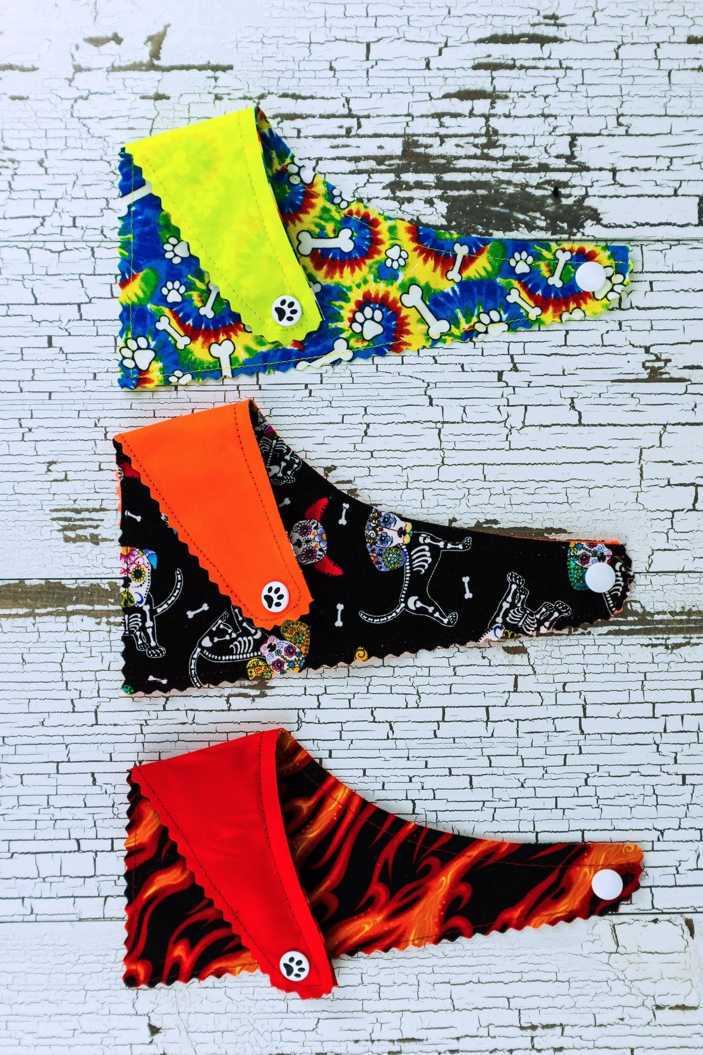 Image shows the reverse side of the reflective bandanas. The neon yellow bandana has a tie dye fabric pattern, the neon orange bandana has a black and white sugar skull dog pattern, and the red bandana has a red and orange flames pattern.