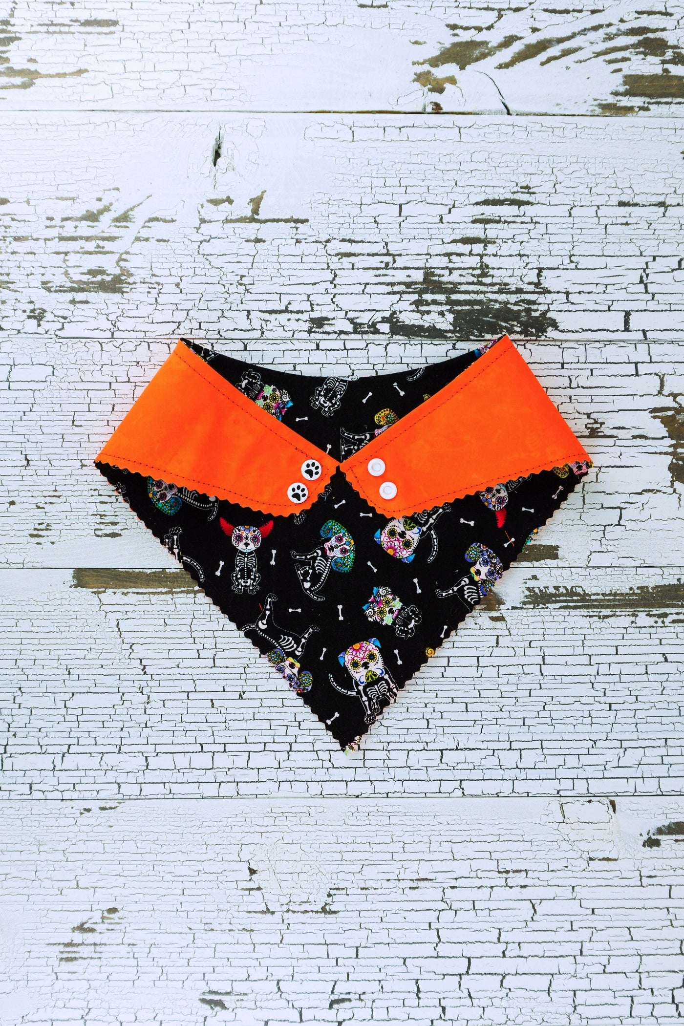 The back of the reversible neon orange bandana is showing the white sugar skull dogs on black fabric. Medium and large bandanas will receive two plastic snaps while the smaller bandanas will get one snap closure.