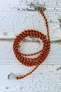 A 15-foot light weight braided orange and black paracord dog training drag line laid out on a white wood background.