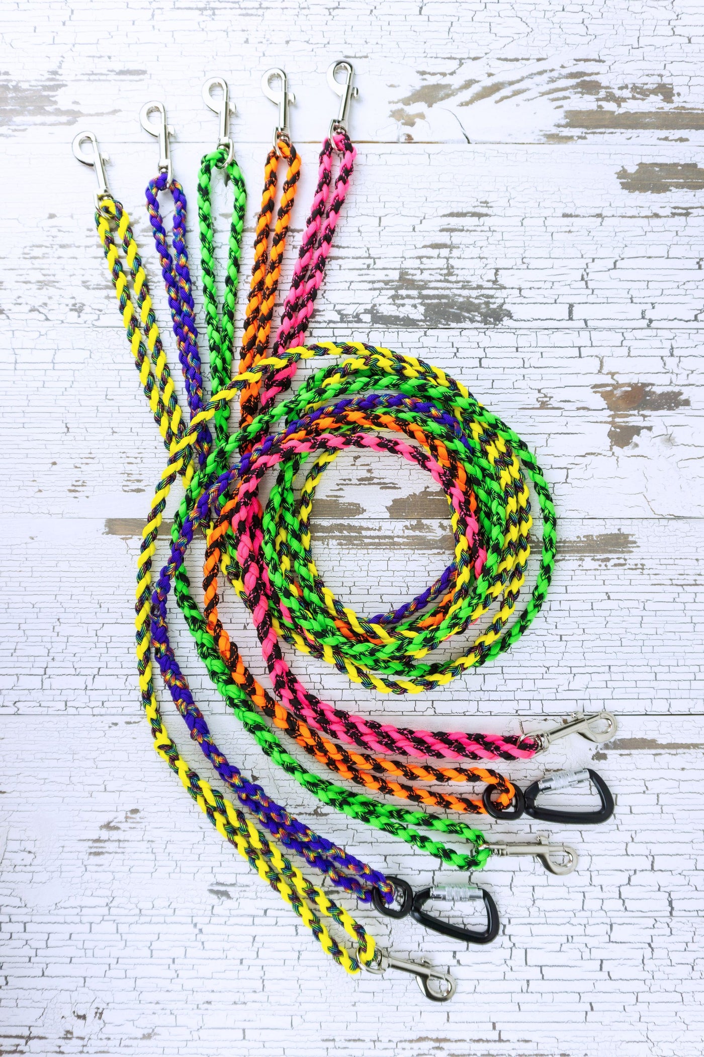 Five different paracord leashes in the two color options of neon yellow and rainbow, purple and rainbow, neon green and black, neon orange and black, and neon pink and black. Leashes are in a swirled flat lay, showing loop handles at each end and two styles of hardware.