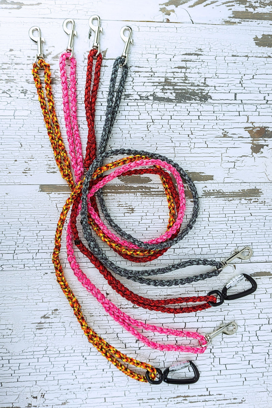 four different paracord leashes in the color blends of orange, pink, red, and gray in a swirled flat lay, showing loop handles at each end and two styles of hardware.