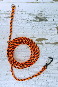 A braided dog drag line with orange and black paracord, featuring a carabiner on one end and o ring on the other, laid out on a white wood background.